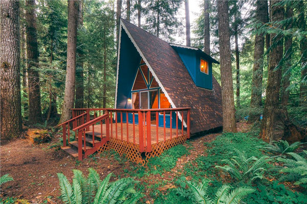 This blue A-frame tiny house with an orange porch and patio for dining looks perfectly appropriate in the forest setting