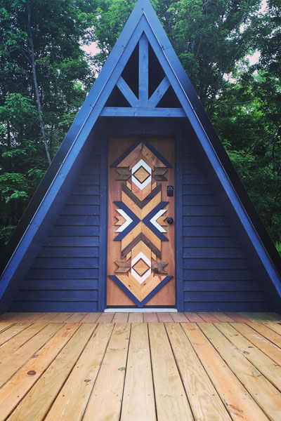 A blue A-frame tiny house looks fantastic with a geometric wooden pattern on the front door.