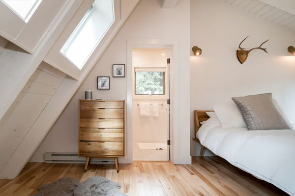 An interior shot of this A-frame tiny house shows a roomy dormered bedroom decorated in white.