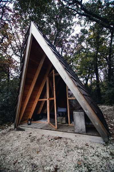 A wooden A-frame tiny house is a nice camping spot on the beach with a porch and windowed front.