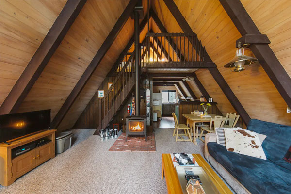An interior of a small A-frame cabin shows how roomy the living room can feel with elevated ceilings and a woodburning stove.
